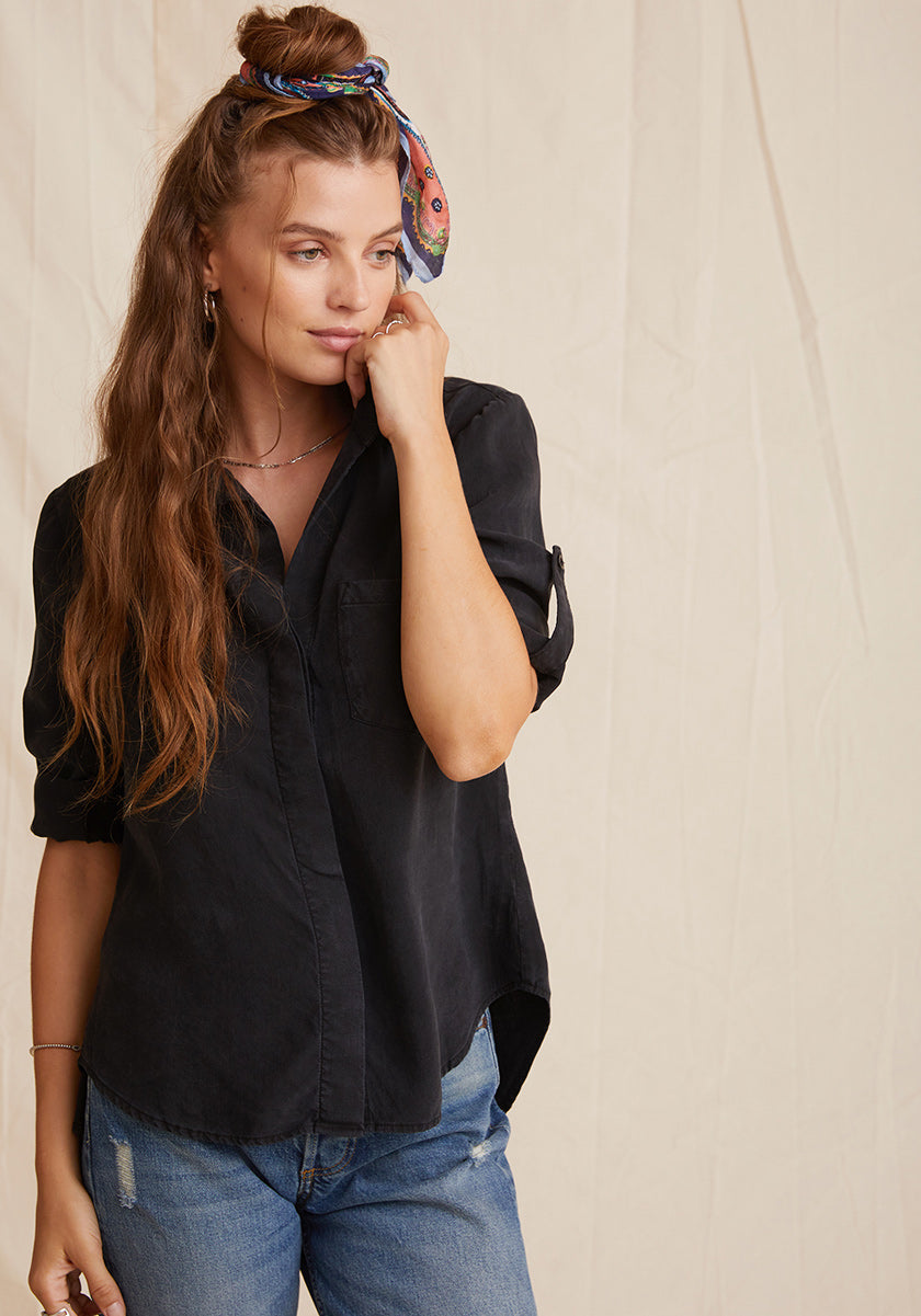 A young woman with long wavy brown hair and a colorful scarf headband stands against a beige backdrop, wearing a loose black Bella Dahl Lyocell-Knopfleiste shirt with rolled sleeves and blue Denimoptik jeans, gazing thoughtfully to the side.