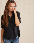 A young woman with long wavy brown hair and a colorful scarf headband stands against a beige backdrop, wearing a loose black Bella Dahl Lyocell-Knopfleiste shirt with rolled sleeves and blue Denimoptik jeans, gazing thoughtfully to the side.