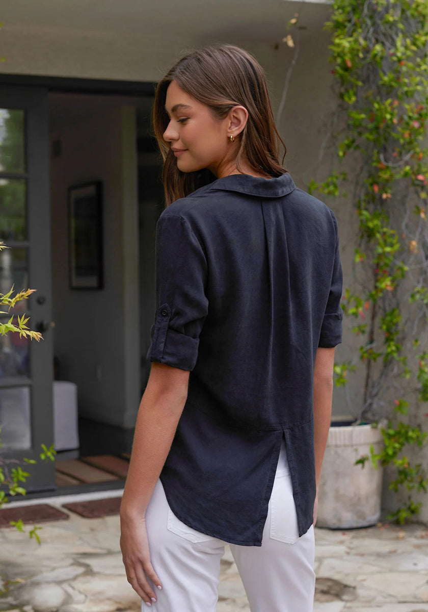 A young woman with straight brown hair stands outdoors near a grey door and green plants, wearing white pants and a Bella Dahl navy blue Lyocell button-up shirt with rolled sleeves, Knopfleiste, and a back slit.