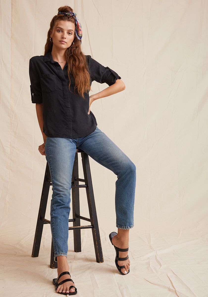 A woman with long brown hair, partly tied up with a colorful scarf, wears a black Bella Dahl Lyocell shirt with Knopfleiste, denim-look blue jeans, and black sandals. She leans on a wooden step stool before a beige, fabric-draped backdrop.