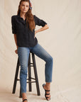 A woman with long brown hair, partly tied up with a colorful scarf, wears a black Bella Dahl Lyocell shirt with Knopfleiste, denim-look blue jeans, and black sandals. She leans on a wooden step stool before a beige, fabric-draped backdrop.