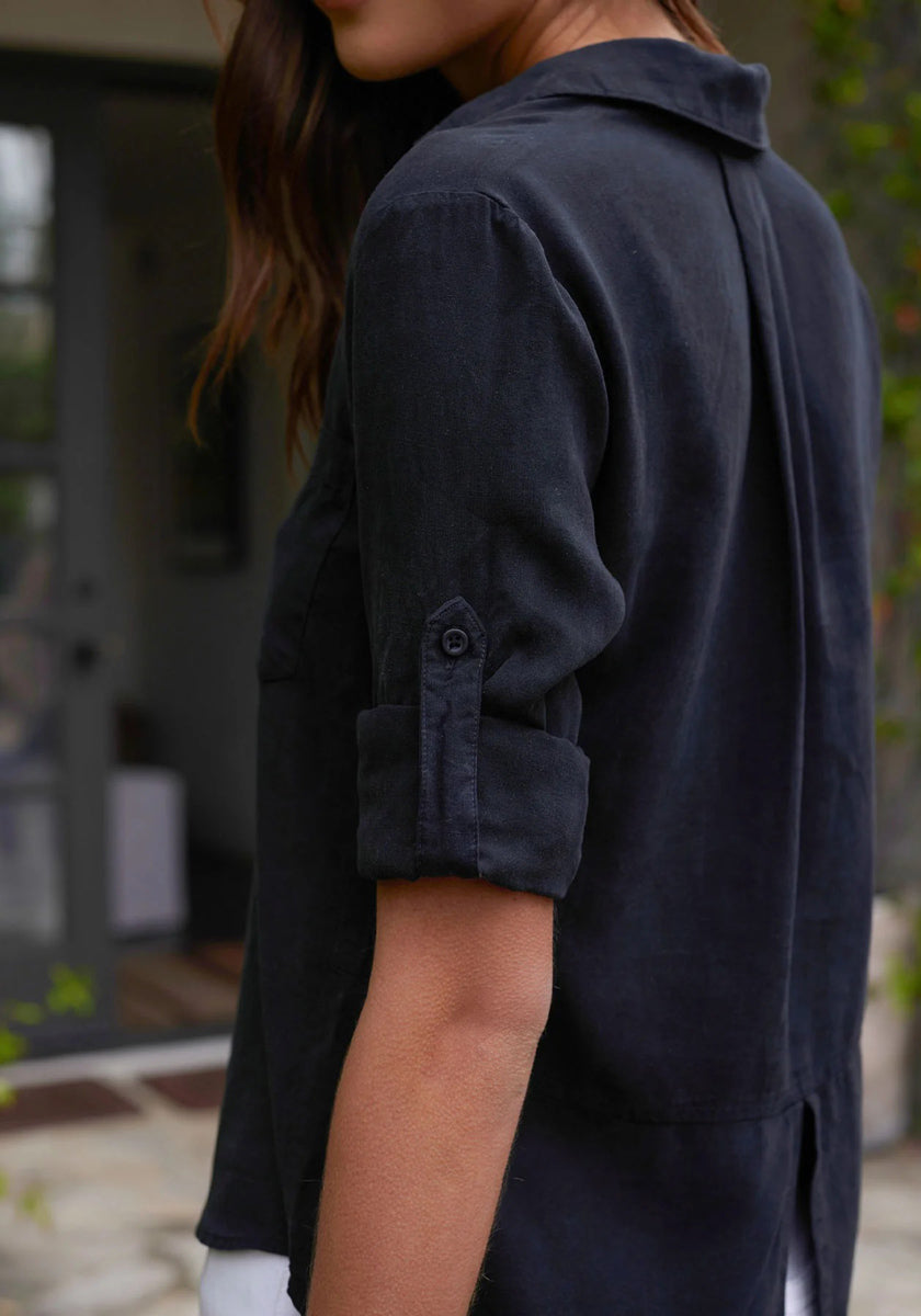 A person with long brown hair wears a dark blue, long-sleeve Bella Dahl shirt in Denimoptik, sleeves rolled up and buttoned at the elbow. The blurred outdoor background features a door, windows, and greenery. Face is not visible.