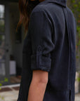 A person with long brown hair wears a dark blue, long-sleeve Bella Dahl shirt in Denimoptik, sleeves rolled up and buttoned at the elbow. The blurred outdoor background features a door, windows, and greenery. Face is not visible.
