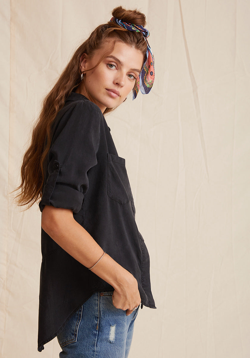 A young woman with long wavy hair wears a Bella Dahl black Lyocell button-up shirt with rolled sleeves, blue denim-look jeans, and a colorful patterned headband. She stands sideways, hand in pocket, against a neutral textured backdrop.