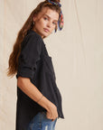 A young woman with long wavy hair wears a Bella Dahl black Lyocell button-up shirt with rolled sleeves, blue denim-look jeans, and a colorful patterned headband. She stands sideways, hand in pocket, against a neutral textured backdrop.