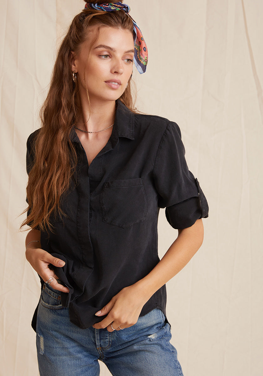 A woman with long, wavy brown hair tied with a colorful scarf stands against a beige fabric backdrop. She wears Bella Dahl’s short-sleeve black Lyocell shirt with Knopfleiste and blue jeans, gazing to the side with one hand in her pocket.