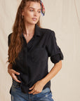 A woman with long, wavy brown hair tied with a colorful scarf stands against a beige fabric backdrop. She wears Bella Dahl’s short-sleeve black Lyocell shirt with Knopfleiste and blue jeans, gazing to the side with one hand in her pocket.