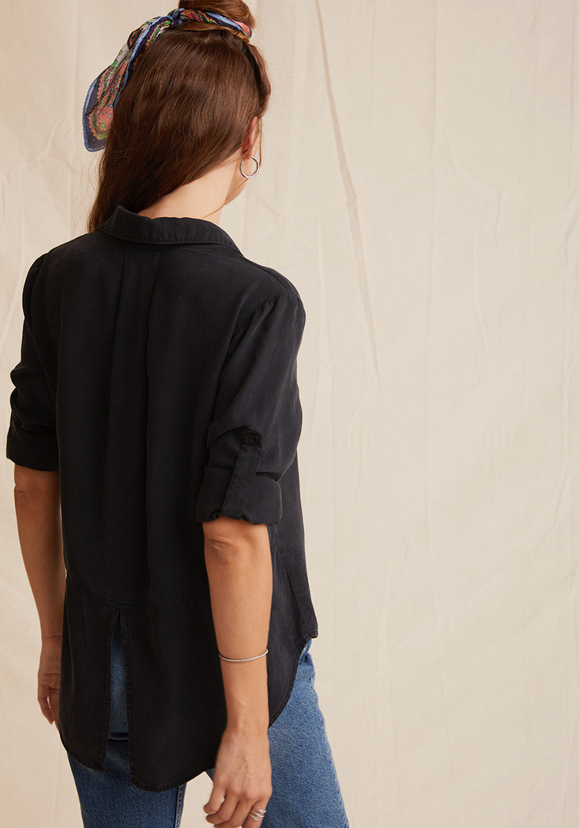 A woman with long brown hair tied with a colorful scarf wears a loose Bella Dahl black long-sleeve shirt with Knopfleiste and blue jeans in Denimoptik as she stands facing away from the camera against a beige fabric backdrop.