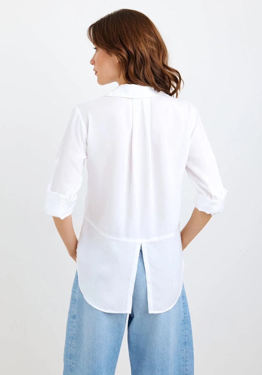A woman with loose, wavy brown hair is seen from behind wearing a long-sleeved Bella Dahl white shirt with rolled-up sleeves, a back pleat, center hem slit, and soft Lyocell fabric, paired with light blue denim-look jeans.
