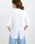 A woman with loose, wavy brown hair is seen from behind wearing a long-sleeved Bella Dahl white shirt with rolled-up sleeves, a back pleat, center hem slit, and soft Lyocell fabric, paired with light blue denim-look jeans.