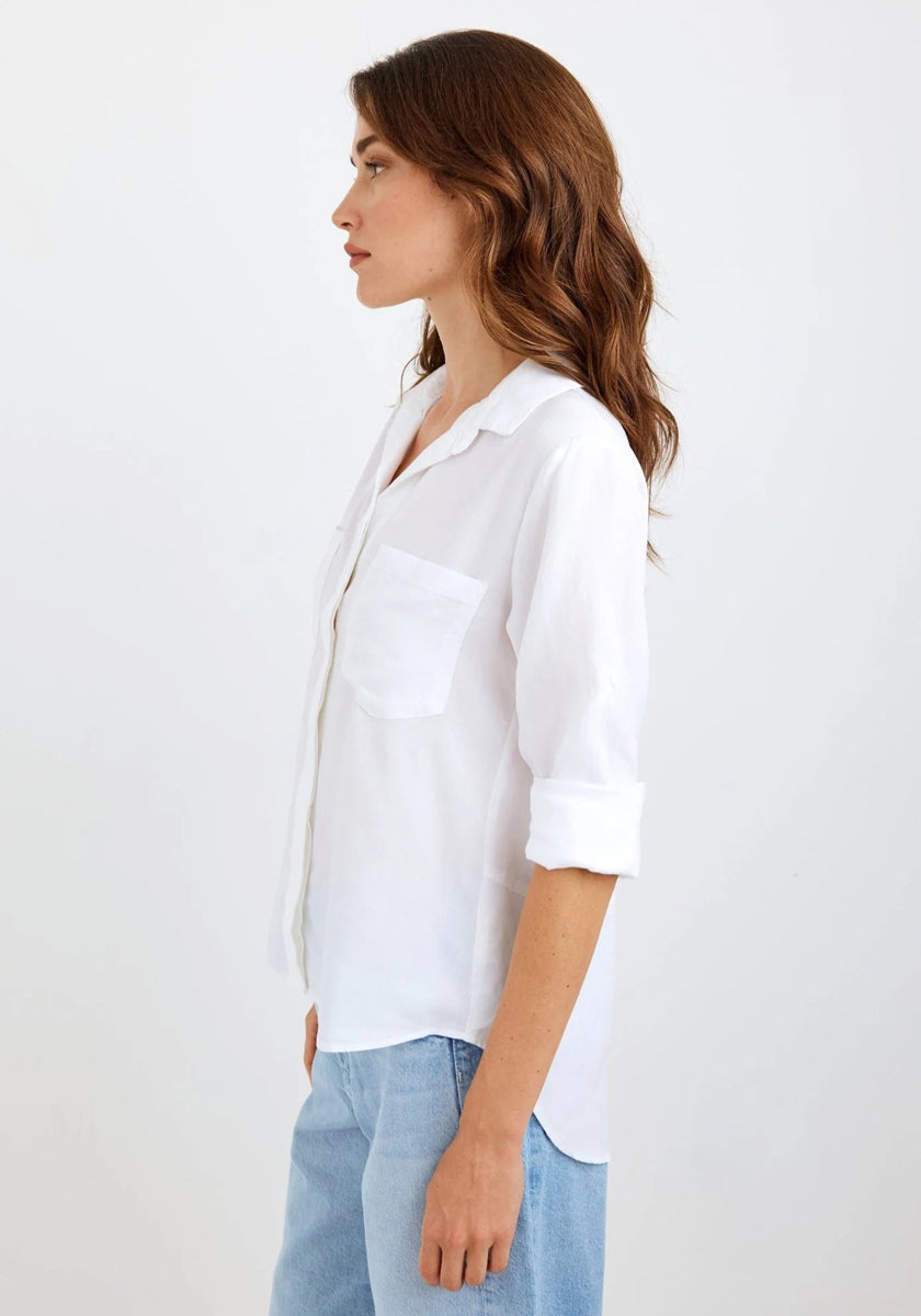 A woman with wavy brown hair stands in profile against a white background, wearing a Bella Dahl white Lyocell Hemd with rolled-up sleeves and a chest pocket, paired with light blue Jeans in Denimoptik. She faces left, her expression neutral.