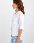 A woman with wavy brown hair stands in profile against a white background, wearing a Bella Dahl white Lyocell Hemd with rolled-up sleeves and a chest pocket, paired with light blue Jeans in Denimoptik. She faces left, her expression neutral.