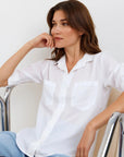 A woman with light skin and brown hair sits on a metal chair, resting her chin on her hand. She wears a loose Bella Dahl Lyocell Hemd with rolled sleeves and blue Jeans in Denimoptik, gazing thoughtfully against a white background.