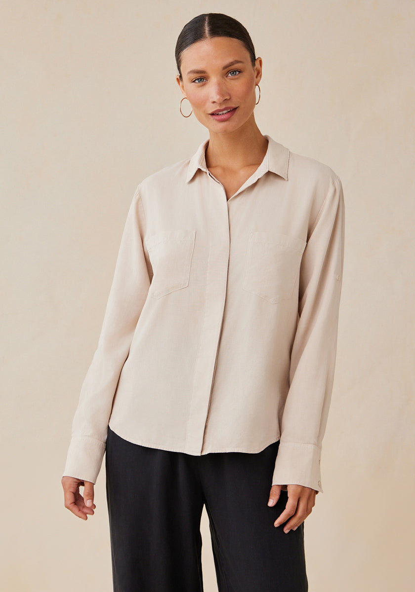 A woman with light skin and slicked-back hair models a loose, light beige Bella Dahl Lyocell button-up shirt with Hemdkragen and long sleeves, paired with black wide-leg pants. She wears large hoop earrings and poses against a plain beige background.
