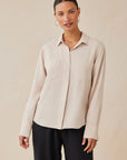 A woman with light skin and slicked-back hair models a loose, light beige Bella Dahl Lyocell button-up shirt with Hemdkragen and long sleeves, paired with black wide-leg pants. She wears large hoop earrings and poses against a plain beige background.