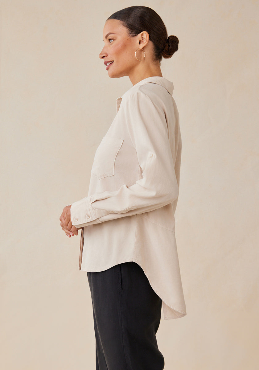 Against a neutral background, a woman with dark hair in a bun wears black pants, gold hoop earrings, and a light beige Bella Dahl Lyocell button-up shirt featuring a Hemdkragen, two breast pockets, and rolled-up sleeves.