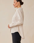 Against a neutral background, a woman with dark hair in a bun wears black pants, gold hoop earrings, and a light beige Bella Dahl Lyocell button-up shirt featuring a Hemdkragen, two breast pockets, and rolled-up sleeves.