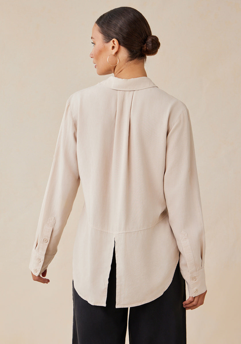 A woman is seen from the back wearing a Bella Dahl light beige long-sleeve blouse with a Hemdkragen, buttoned cuffs, and a back slit at the hem. Her hair is in a low bun, paired with large hoop earrings and loose black pants.