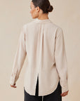 A woman is seen from the back wearing a Bella Dahl light beige long-sleeve blouse with a Hemdkragen, buttoned cuffs, and a back slit at the hem. Her hair is in a low bun, paired with large hoop earrings and loose black pants.