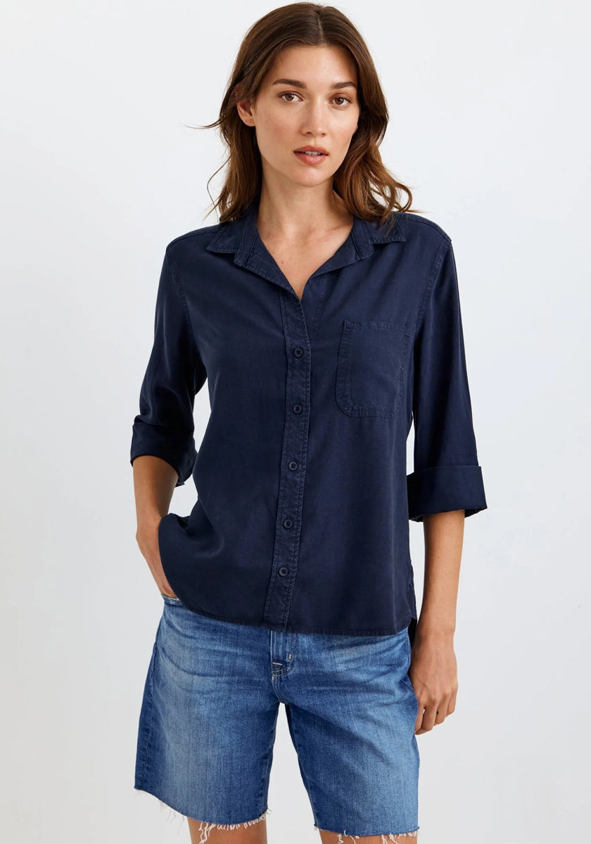 A woman with long brown hair stands in a relaxed pose against a plain light background, wearing an oversized Bella Dahl navy blue Lyocell shirt with rolled sleeves and a chest pocket, paired with knee-length blue denim shorts. One hand is in her pocket.
