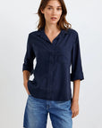 A woman with long brown hair stands in a relaxed pose against a plain light background, wearing an oversized Bella Dahl navy blue Lyocell shirt with rolled sleeves and a chest pocket, paired with knee-length blue denim shorts. One hand is in her pocket.