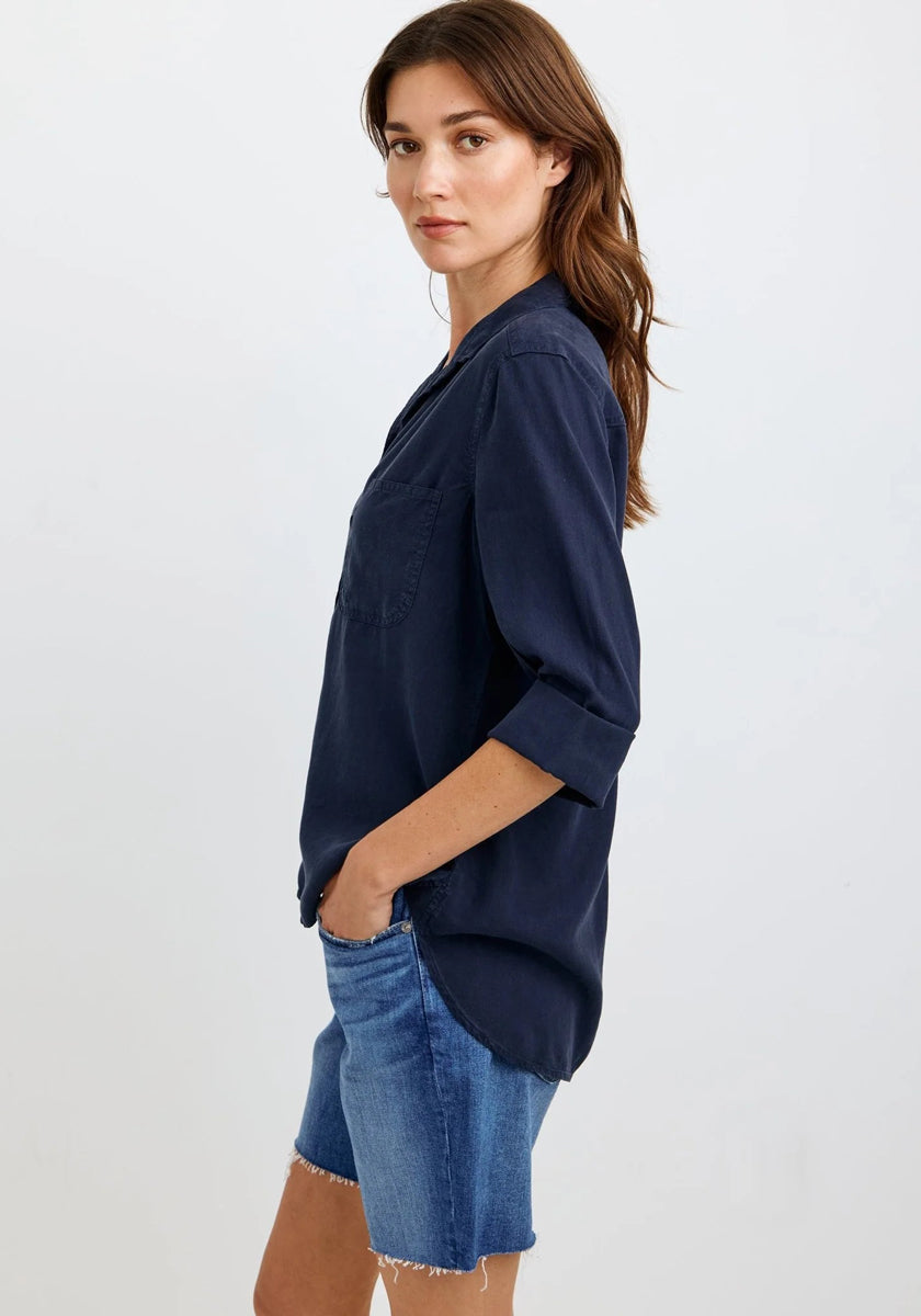 A woman with long brown hair stands sideways, looking at the camera. She wears a navy Bella Dahl Oversized Schnitt Hemdbluse with sleeves rolled up and blue frayed denim shorts. Her left hand is in her pocket; the background is light-colored.