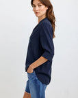 A woman with long brown hair stands sideways, looking at the camera. She wears a navy Bella Dahl Oversized Schnitt Hemdbluse with sleeves rolled up and blue frayed denim shorts. Her left hand is in her pocket; the background is light-colored.