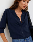 A woman with long brown hair wears a navy oversized Hemdbluse by Bella Dahl, its top buttons undone and sleeves rolled up. She pairs it with blue jeans and a black woven bag, standing against a plain white wall and looking downward.
