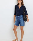 A woman in a relaxed pose stands by a white wall, wearing a navy Bella Dahl Lyocell Hemdbluse with an oversized cut and rolled sleeves, paired with mid-length blue raw-hem denim shorts, white flat sandals, and a black textured shoulder bag.