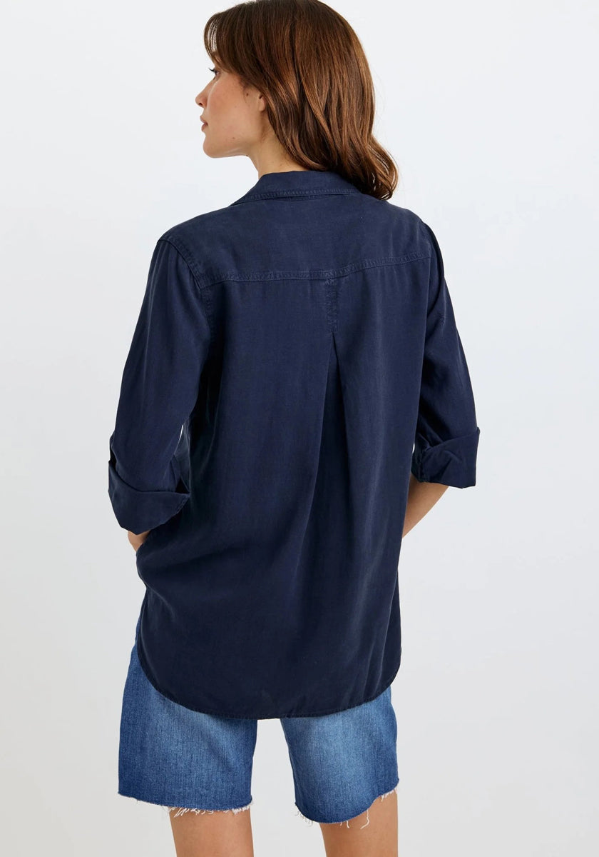 A woman with brown hair faces away from the camera, wearing an oversized Bella Dahl dark navy Lyocell hemdbluse with rolled-up sleeves and loose faded blue denim shorts against a plain white background.