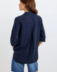 A woman with brown hair faces away from the camera, wearing an oversized Bella Dahl dark navy Lyocell hemdbluse with rolled-up sleeves and loose faded blue denim shorts against a plain white background.