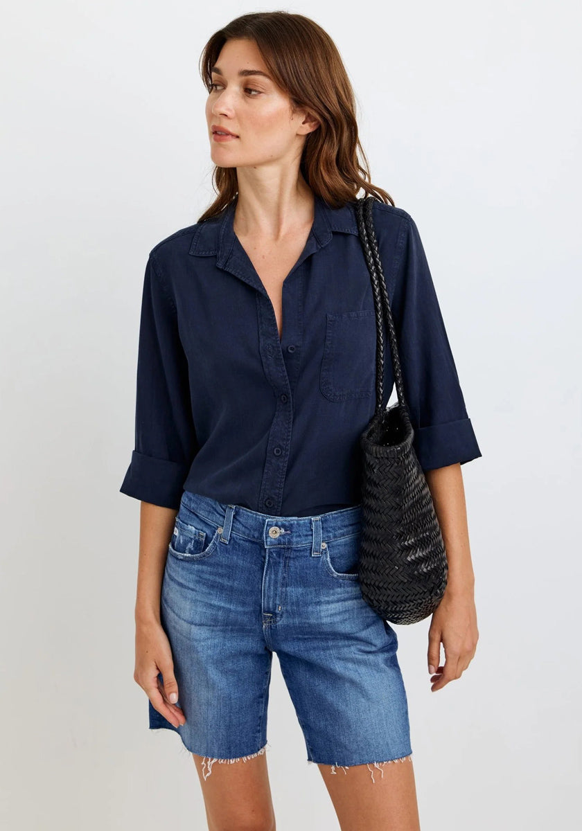A woman with wavy brown hair wears a navy oversized Hemdbluse by Bella Dahl with rolled sleeves and blue frayed denim shorts. She carries a black woven shoulder bag, stands against a light background, and looks left with a relaxed expression.