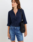 A woman with wavy brown hair wears a navy oversized Hemdbluse by Bella Dahl with rolled sleeves and blue frayed denim shorts. She carries a black woven shoulder bag, stands against a light background, and looks left with a relaxed expression.