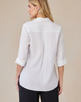 A woman with long blonde hair is seen from behind, wearing an oversized white Lyocell shirt by Bella Dahl with rolled-up sleeves over black pants, set against a simple light beige background.