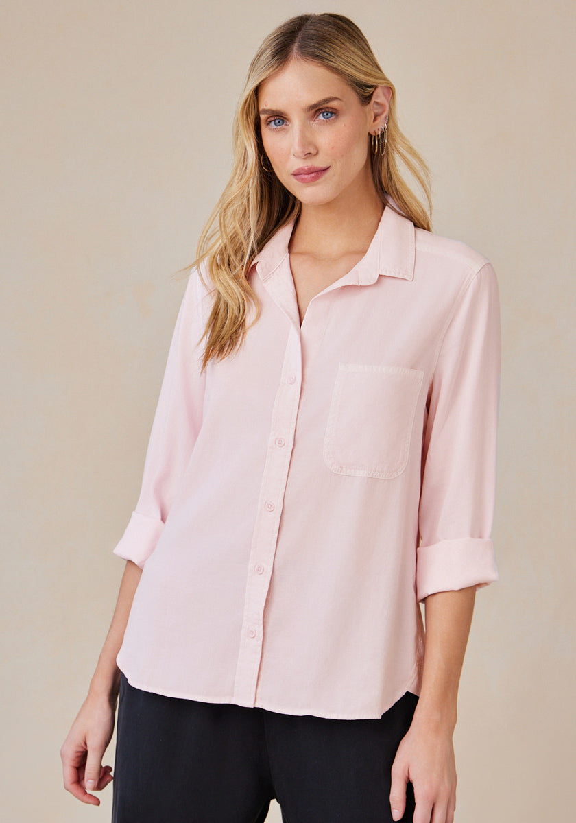 A woman with long blonde hair wears a light pink Bella Dahl Lyocell Hemdbluse with a front pocket and rolled sleeves, paired with black pants and small hoop earrings, standing against a beige background and smiling slightly at the camera.