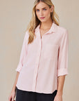A woman with long blonde hair wears a light pink Bella Dahl Lyocell Hemdbluse with a front pocket and rolled sleeves, paired with black pants and small hoop earrings, standing against a beige background and smiling slightly at the camera.