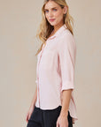 A woman with long blonde hair stands in profile, wearing a light pink oversized Hemdbluse by Bella Dahl with rolled sleeves and a front pocket, paired with dark pants. She poses against a plain beige background with a neutral, pleasant expression.