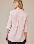 A woman with long blonde hair stands facing away, wearing a pale pink Bella Dahl Lyocell Hemdbluse with rolled-up sleeves, pleat in the back, oversized Schnitt, and relaxed fit, paired with black pants against a beige backdrop.
