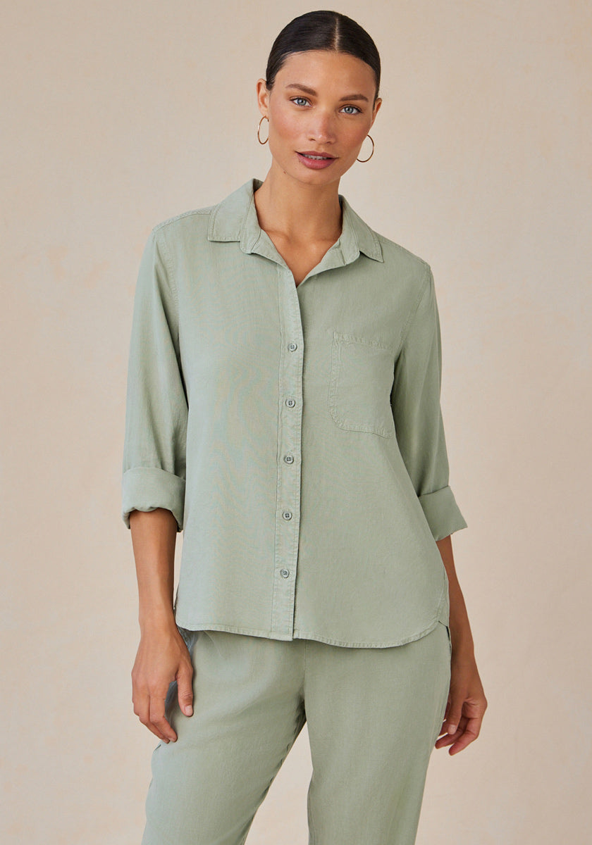 A woman with dark hair tied back wears a sage green Bella Dahl Lyocell Hemdbluse with Knopfleiste, rolled sleeves, and a chest pocket, paired with matching pants. She stands against a plain beige background, looking forward neutrally.
