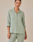 A woman with dark hair tied back wears a sage green Bella Dahl Lyocell Hemdbluse with Knopfleiste, rolled sleeves, and a chest pocket, paired with matching pants. She stands against a plain beige background, looking forward neutrally.