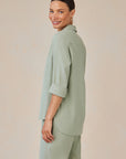 A woman with medium skin and dark hair tied back smiles over her shoulder. She wears a light sage green Lyocell Hemdbluse with button placket and rolled sleeves by Bella Dahl, paired with matching wide-leg pants against a neutral beige background.