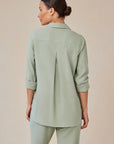 A woman with dark hair in a low bun faces away, wearing a light sage green Bella Dahl Lyocell Hemdbluse with rolled-up sleeves and matching pants. The shirt has a back pleat, relaxed fit, subtle Knopfleiste, set against a light beige background.