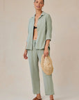 A woman smiles in a relaxed light sage green Bella Dahl Lyocell Hemdbluse with Knopfleiste over a black bralette, paired with matching drawstring pants, brown sandals, large hoop earrings, and a woven straw bag. Her hair is pulled back against a plain beige backdrop.