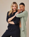 Two women pose against a light beige background. One wears a black oversized Hemdbluse and pants, the other sports a sage green Bella Dahl shirt with matching Lyocell pants. Smiling, they link arms as one leans on the others shoulder.