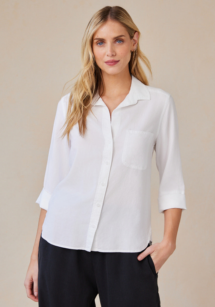 A woman with long blonde hair wears a white Bella Dahl Hemdbluse aus 100% Lyocell with a front pocket and 3/4 sleeves, standing against a beige background, one hand in her black pants pocket, highlighting the oversized Schnitt.