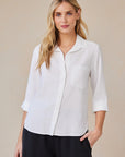 A woman with long blonde hair wears a white Bella Dahl Hemdbluse aus 100% Lyocell with a front pocket and 3/4 sleeves, standing against a beige background, one hand in her black pants pocket, highlighting the oversized Schnitt.