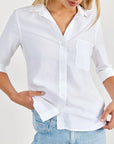 A woman in a crisp white Bella Dahl Hemdbluse with chest pocket and rolled-up sleeves, styled oversized, pairs it with light blue jeans. Her blonde hair falls loosely as she slips a hand into her pocket. The shirt is made from 100% Lyocell.