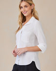 A woman with long blonde hair smiles in a white Bella Dahl 100% Lyocell Hemdbluse with chest pocket, oversized fit, and 3/4 rolled sleeves, styled with hoop earrings and black pants against a beige background.