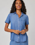 A woman with long, wavy hair wears a Bella Dahl short-sleeve lyocell blouse with shirt collar and chest pocket in light blue denim. She stands against a plain light gray background, smiling slightly with her hands at her waist.
