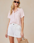 A woman with long blonde hair wears sunglasses, a light pink Lyocell blouse with a shirt collar by Bella Dahl, and high-waisted white shorts. She holds a small brown woven handbag. The background is neutral beige.
